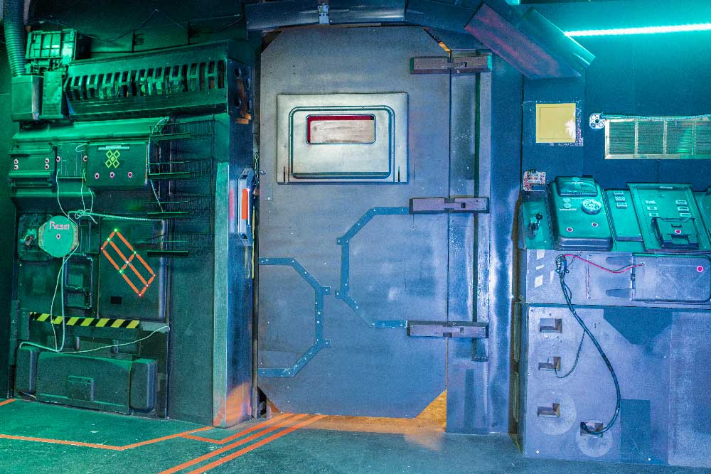 Wide shot of a sci-fi escape room interior featuring a heavy metal door, control panels, and atmospheric green lighting at Escape Ops Calgary
